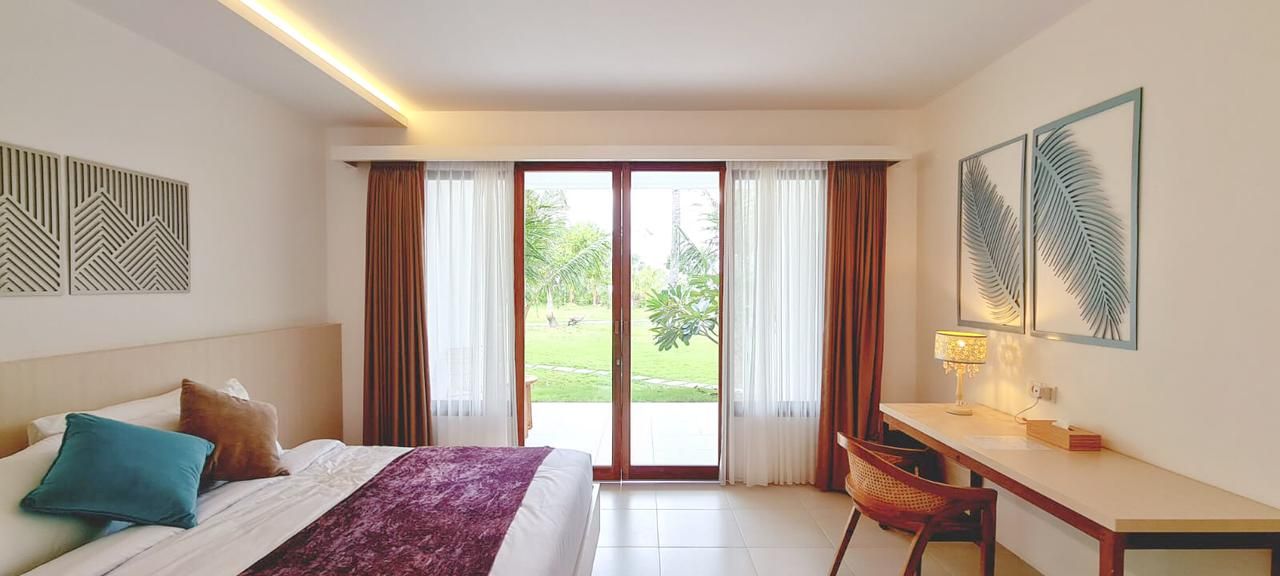 A hotel room with a bed, desk, chair, and large sliding glass doors leading to a garden.