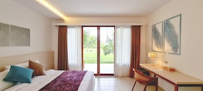 A hotel room with a bed, desk, chair, and large sliding glass doors leading to a garden. - Palm Springs Bali Dive Resorts (Tulamben)
