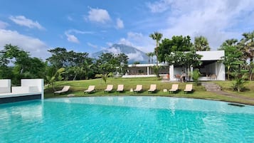 Una piscina con sillas de descanso, un edificio con zona cubierta y una montaña al fondo.