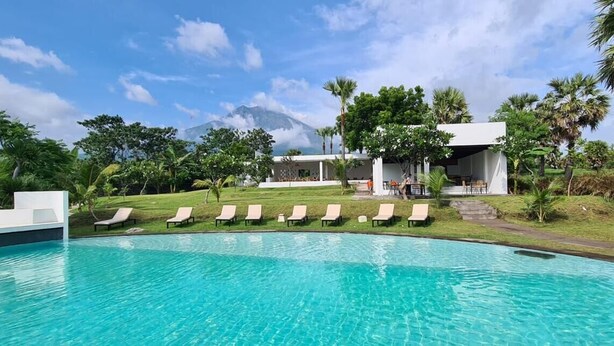 Ein Schwimmbecken mit Liegestühlen, ein Gebäude mit überdachtem Bereich und ein Berg im Hintergrund.