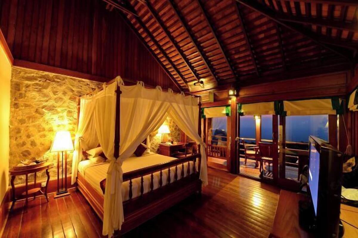 A room with a four-poster bed, stone walls, wooden flooring, and a balcony with a view.