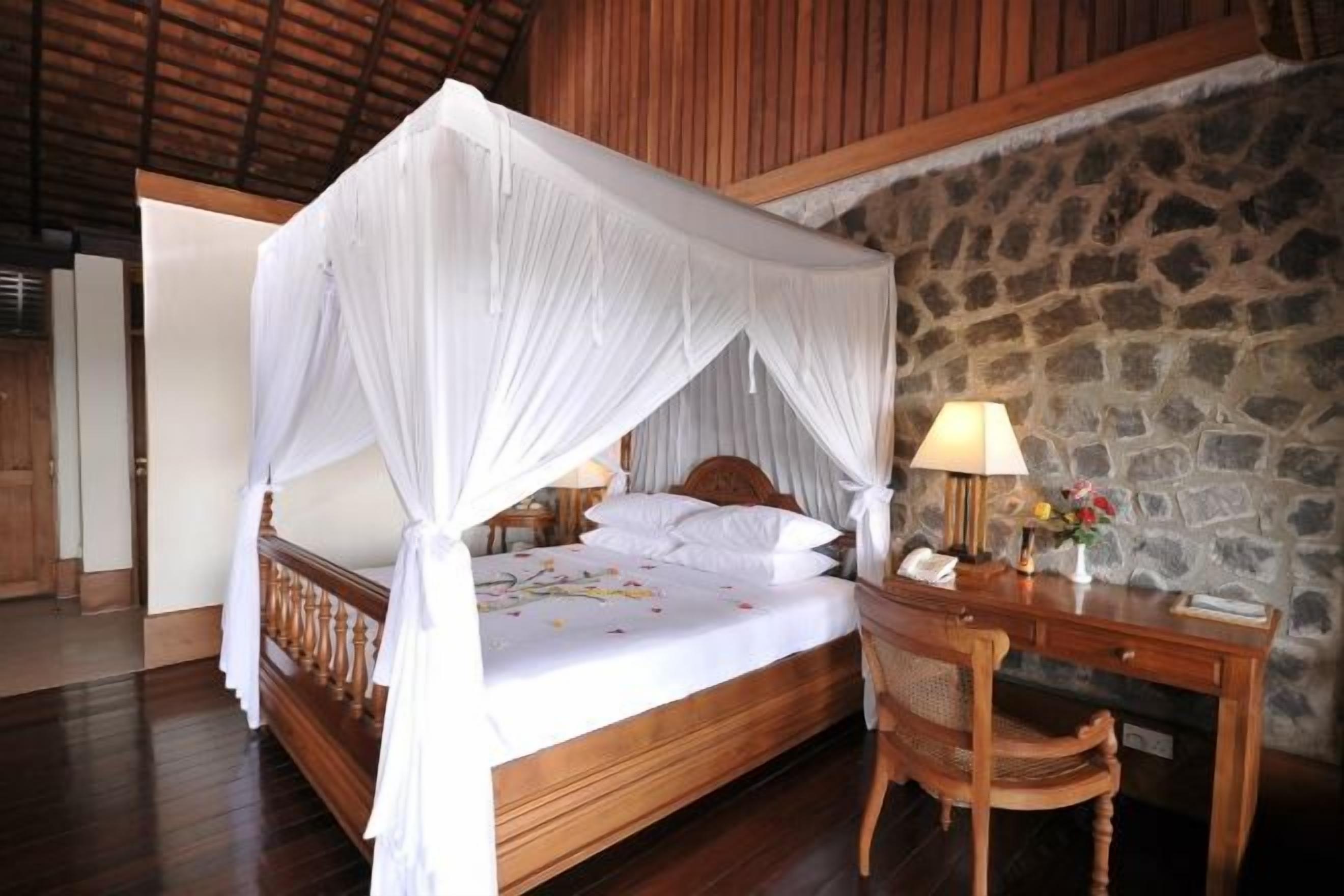 A bed with a canopy, a desk with a lamp, and a stone wall.