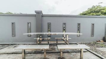 Terraço no telhado com mesa e bancos de metal, parede cinza com janelas, e céu nublado.