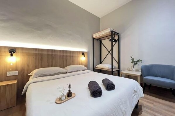 Una habitación de hotel con una cama, mesitas de noche, un sillón azul y un cabecero de madera.
