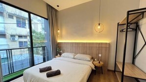 A modern bedroom with a large bed, a bedside table, a hanging light, and a view of buildings through a window.