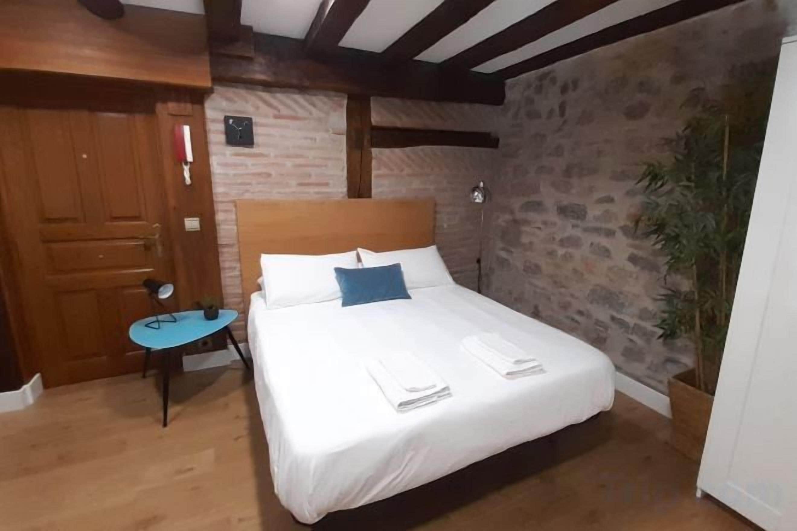 A bedroom with a bed, a wooden door, a small table, and a potted plant.