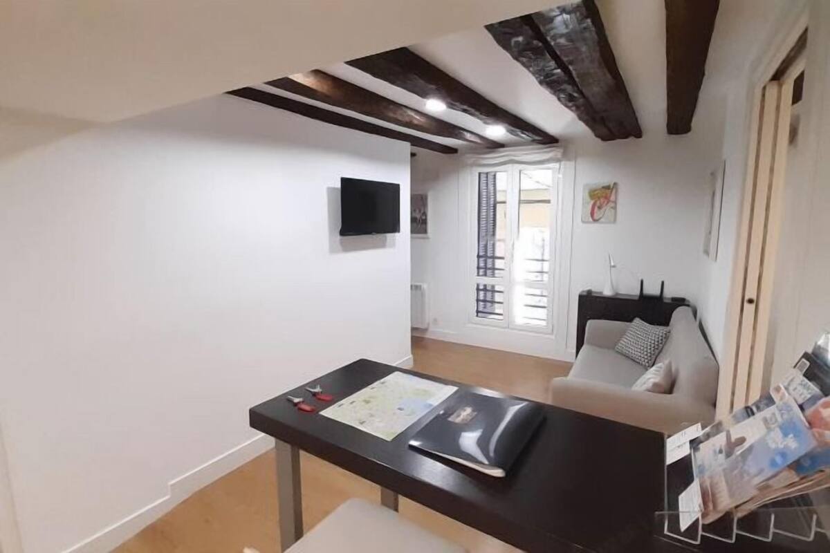 Quarto com teto de madeira, parede branca, mesa preta com papéis, sofá branco e janela com persianas.