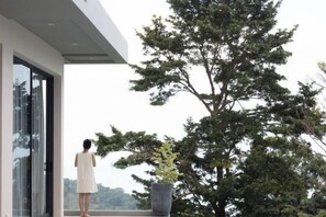 Una persona parada junto a un ventanal grande con vistas a un árbol y al cielo.