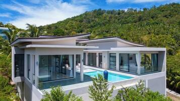 A modern house with a glass-enclosed swimming pool, surrounded by lush greenery and hills.