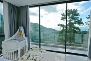 A room with a crib, a large window, and a view of a hilly landscape. - THE PURE BANGPOR (Koh Samui)