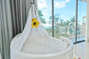 A white crib with a white canopy and a yellow flower decoration, positioned near a window with a view of palm trees and buildings. - THE PURE BANGPOR (Koh Samui)