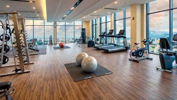 A spacious gym with cardio machines, weightlifting equipment, and large windows offering a city view. - Luxury Ocean View Suites_My Khe Beach_Hinata Apartment (Da Nang)