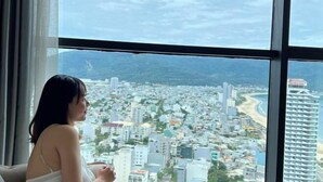 A view from a high-rise hotel window overlooking a coastal cityscape with buildings and a beach. - Luxury Ocean View Suites_My Khe Beach_Hinata Apartment (Da Nang)