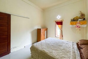 A bedroom with a bed, a wooden wardrobe, a window with curtains, and a wall-mounted shelf with decorative items.