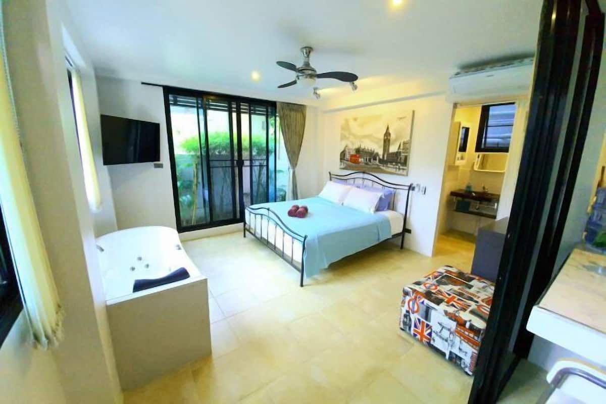 A bedroom with a bed, a ceiling fan, a window with curtains, a small table, and a TV mounted on the wall.