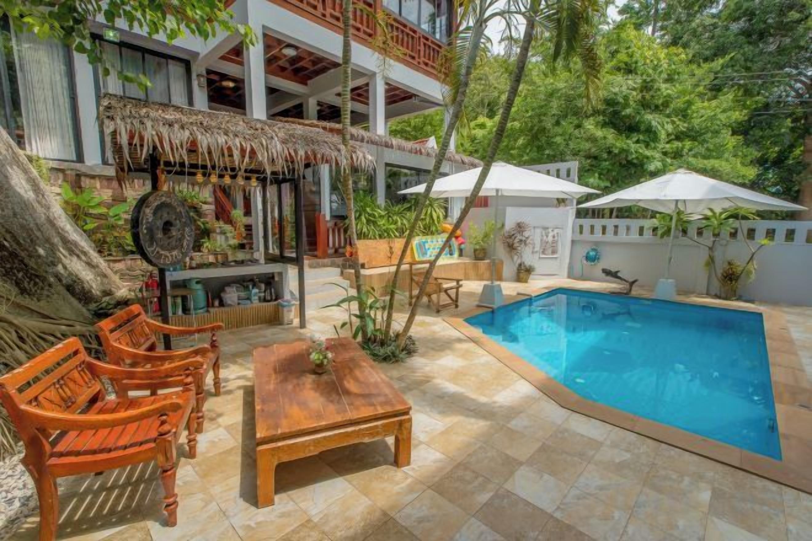 A pool area with a wooden table and chairs, a bar area with a thatched roof, and a white umbrella.