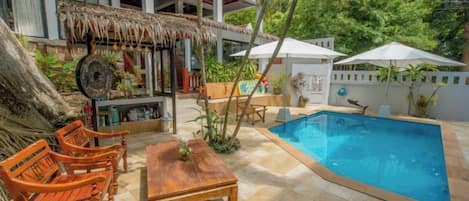 A pool area with a wooden table and chairs, a bar area with a thatched roof, and a white umbrella.