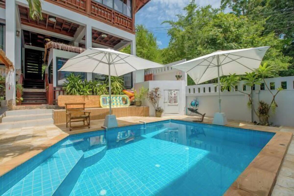 A swimming pool with two umbrellas, a wooden bench, and a small table.