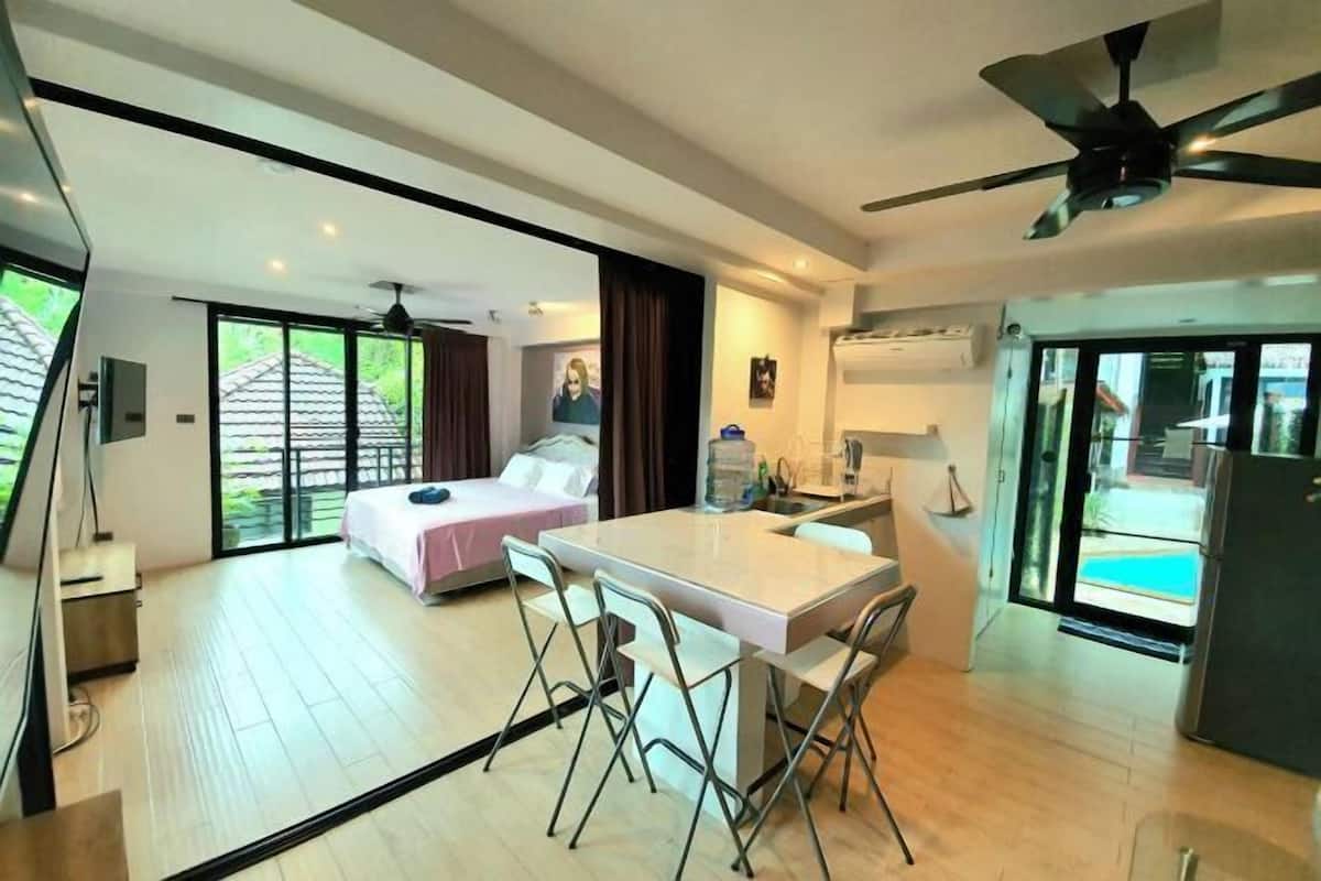 A modern room with a bed, a dining table with chairs, a ceiling fan, and a view of a swimming pool.