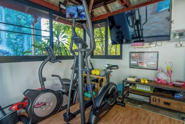 A home gym with exercise equipment, a television, and a window with a view of trees. - Penthouse Crystal Bay Apartments (Koh Samui)