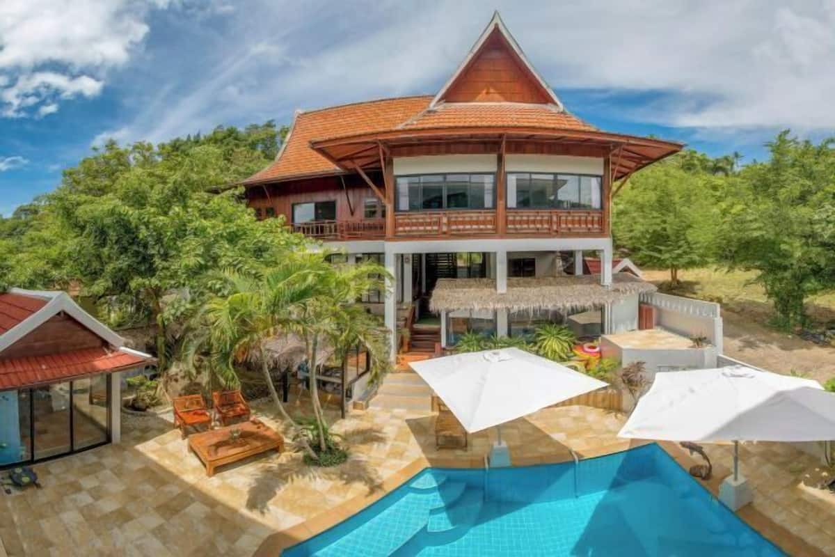 A two-story house with a pool, surrounded by greenery and a thatched-roof structure.