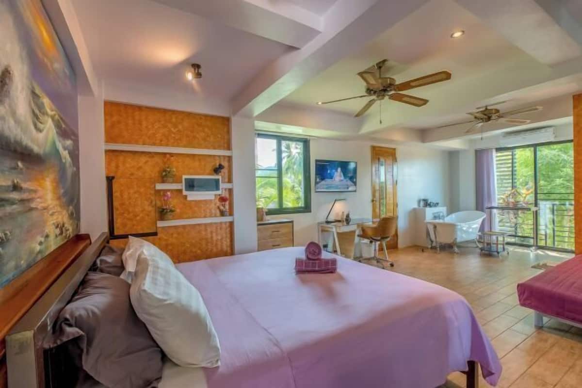 A bedroom with a bed, a desk, a chair, a bathtub, and a balcony with a view of greenery.