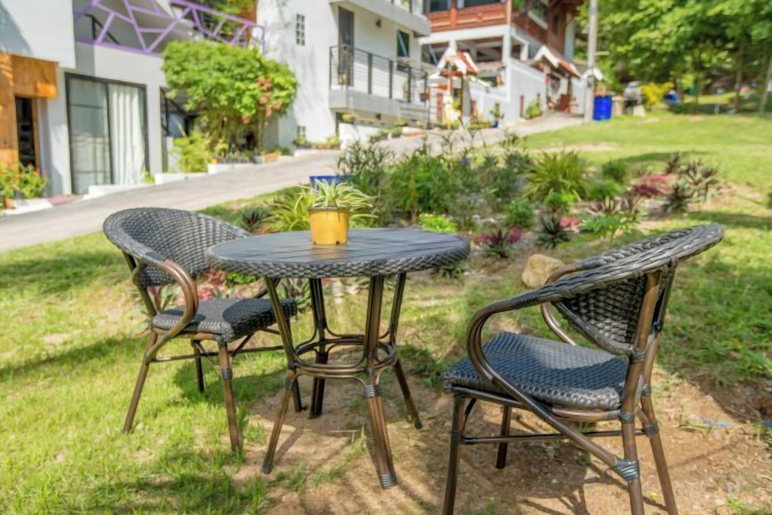 Un giardino con un tavolo rotondo e sedie in vimini, immerso nel verde e con un sentiero che conduce a un edificio.