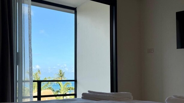 A hotel room with a bed, a window with a view of palm trees and the ocean, and a black-framed window on the wall. - Sman Resort (Koh Samui)