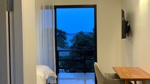 A room with a bed, a wooden table, a chair, a TV, and a view of a body of water through a large window. - Sman Resort (Koh Samui)