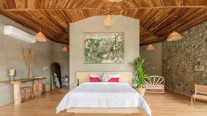 Een slaapkamer met een houten plafond, een bed met witte beddengoed, een stenen muur en een schilderij van een tropisch landschap.