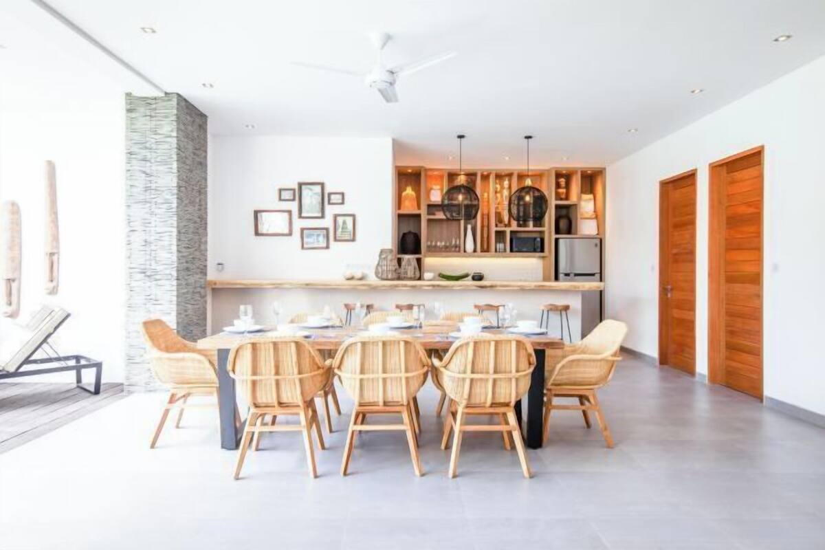 A modern dining area with a long table and six chairs, a kitchen with wooden cabinets and pendant lights, and a stone accent wall.