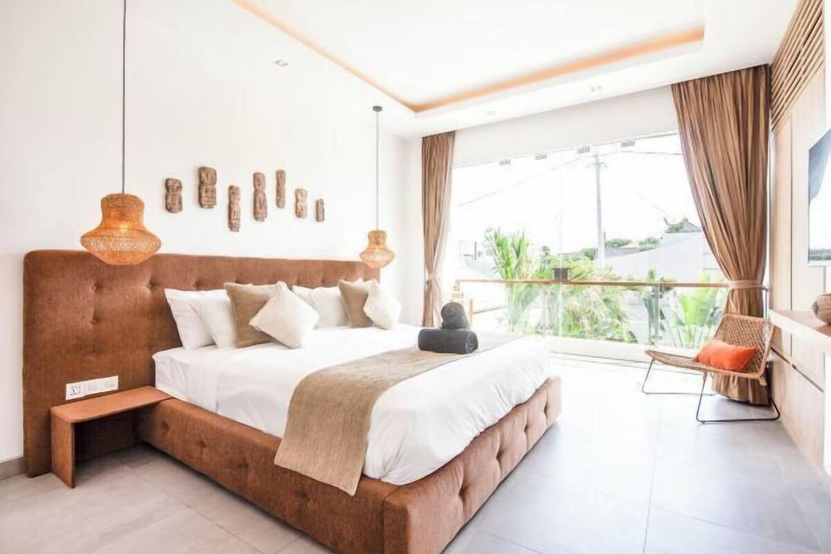 A modern bedroom with a large bed, a small bedside table, a chair, and a balcony with a view.
