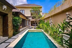 Una piscina rectangular con borde de piedra, rodeada de abundante vegetación y estatuas.