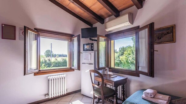 Une pièce avec un plafond en bois, un bureau avec une chaise, un lit recouvert d’une couverture verte et deux fenêtres donnant sur un paysage verdoyant.