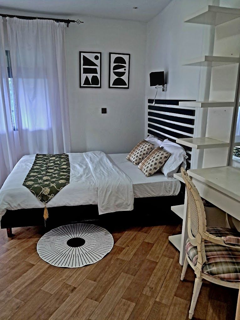 Een slaapkamer met een bed, een rond bijzettafeltje, een stoel en een bureau. Boven het bed hangen twee decoratieve schilderijen.