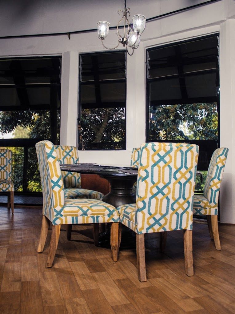 Un comedor con una mesa redonda y cuatro sillas de diseño geométrico, ventanas amplias con vistas a árboles y una lámpara de araña.