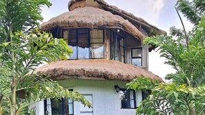 A two-story house with a thatched roof, surrounded by lush greenery.