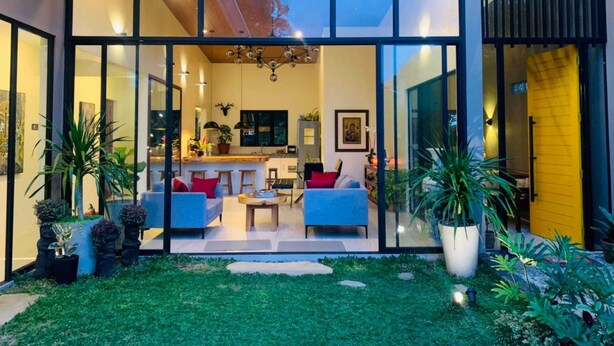 A modern house with a glass facade, a dining area with a blue sofa and a small table, and a kitchen visible through the glass. - OYO 1177 The Great Molave (Alfonso)