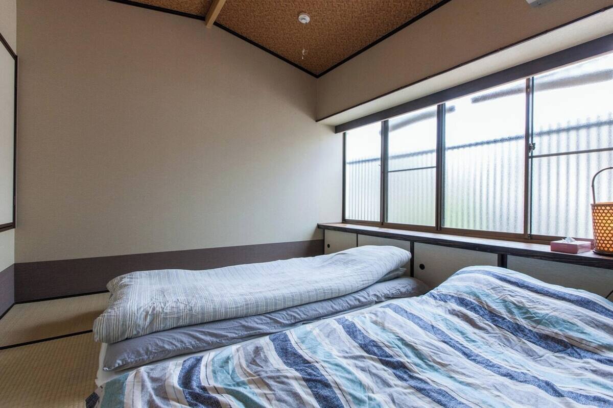 Une chambre avec deux lits, une fenêtre avec des stores, un ventilateur de plafond et une table de chevet avec une lampe.