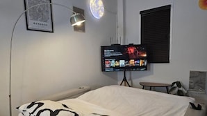 A bedroom with a bed, a TV mounted on the wall, a small table, and a window with blinds. - W. House for group, Cozy Loft near Suvarnabhumi Airport (Bangkok)