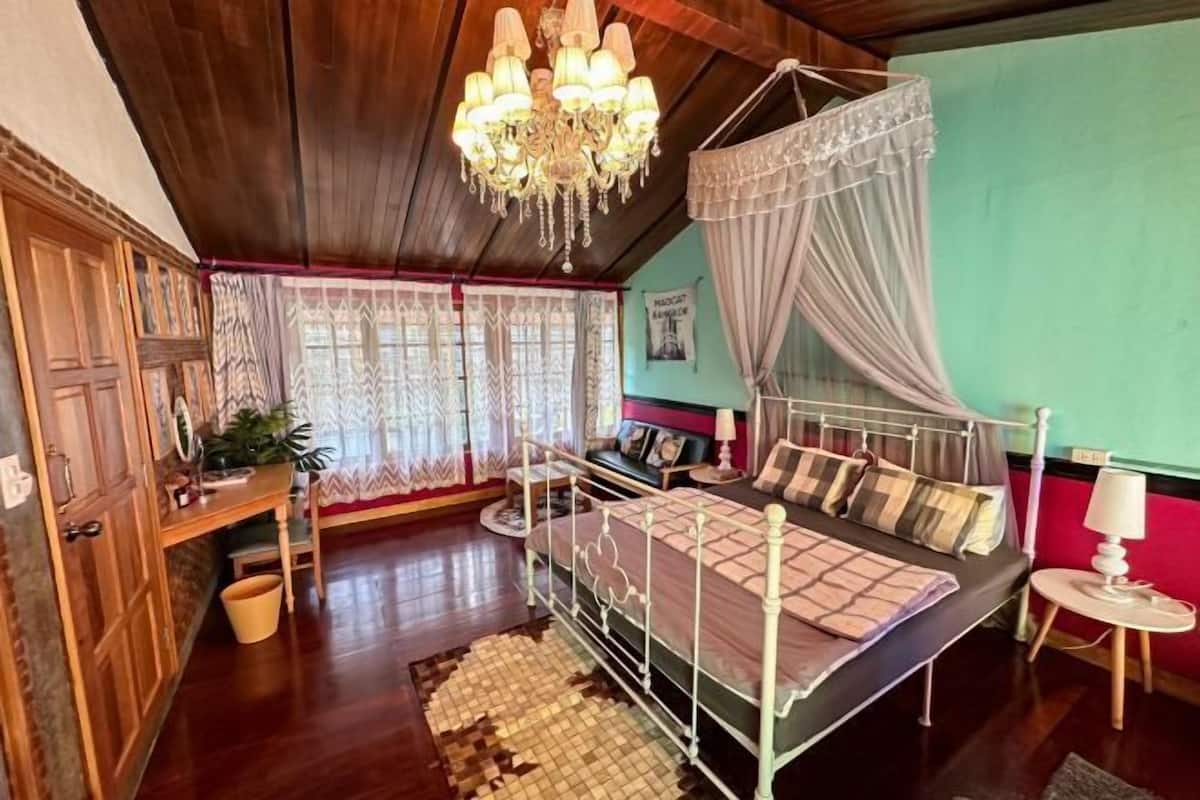 A bedroom with a four-poster bed, wooden flooring, a chandelier, and a window with lace curtains.