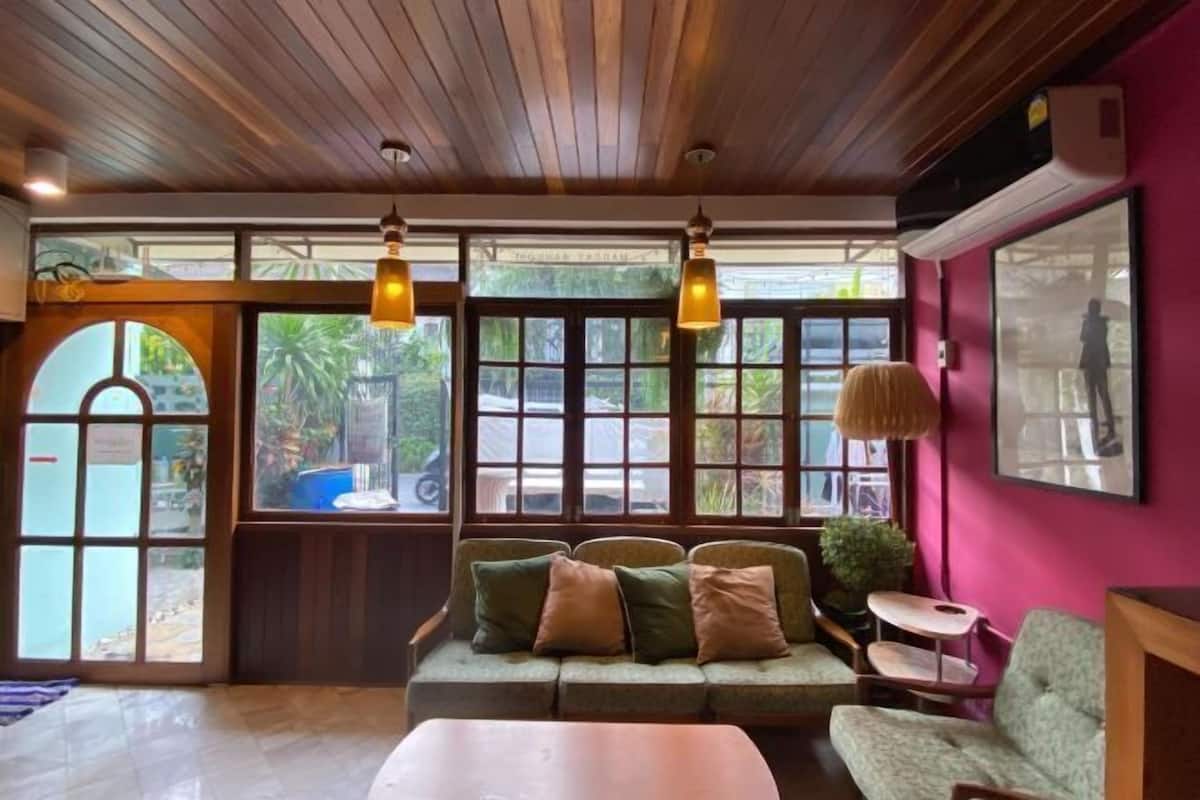 A cozy room with a wooden ceiling, a green sofa with cushions, a small table, and a window with a view of greenery outside.