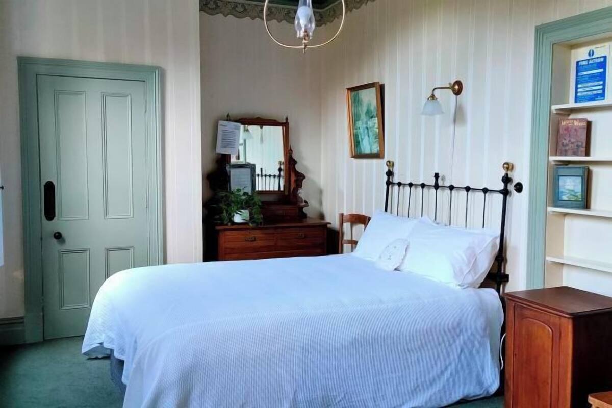 A bedroom with a bed, dresser, mirror, wall-mounted lamp, and a door.