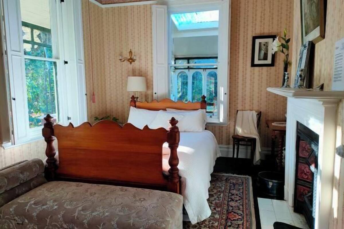 A bedroom with a wooden bed, a bedside table, a chair, a fireplace, and a window with a view of greenery.