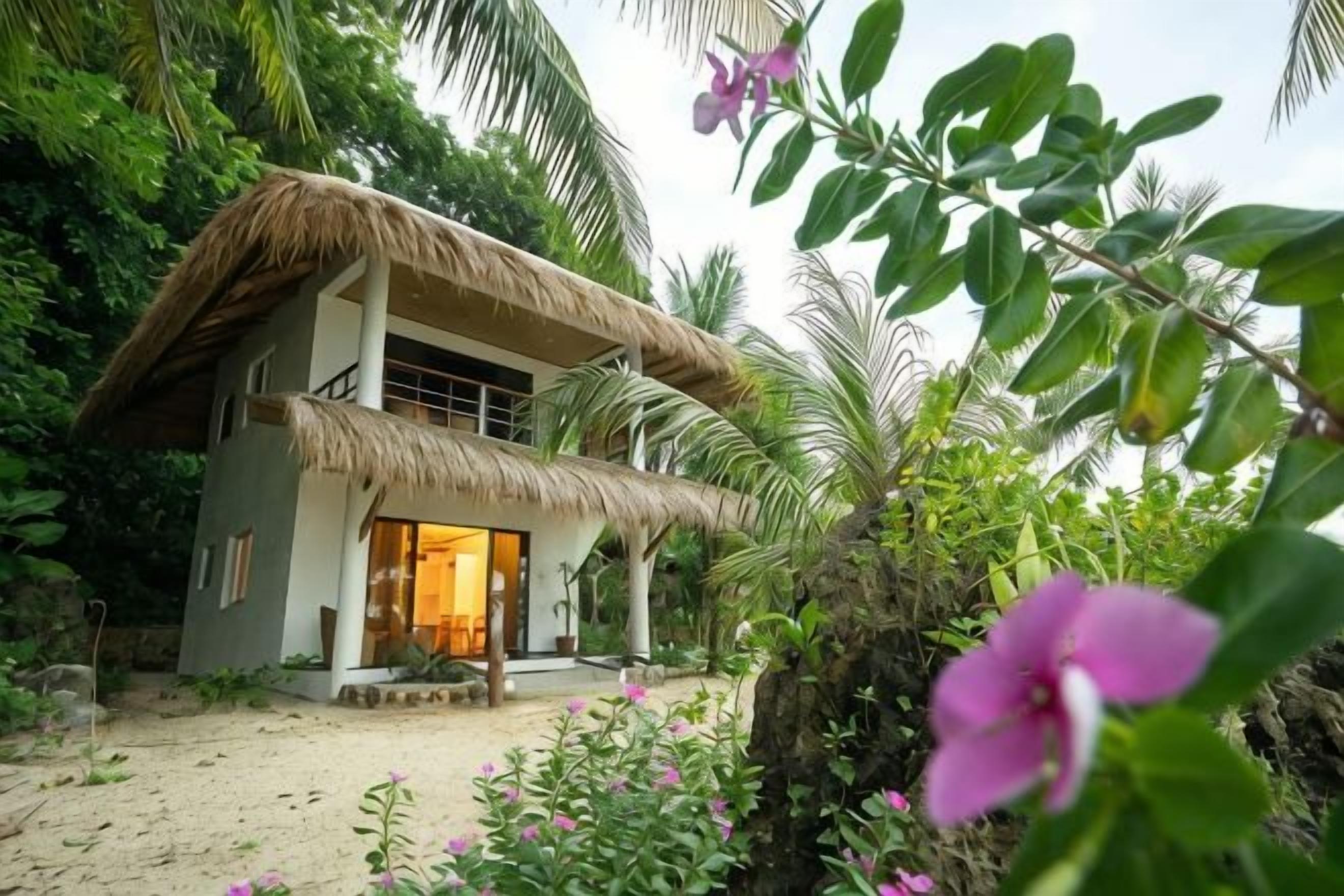 A two-story house with a thatched roof, surrounded by tropical vegetation.