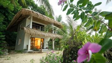 A two-story house with a thatched roof, surrounded by tropical vegetation.