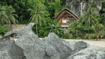 Eine Holzhütte mit einem Reetdach, umgeben von Palmen und Felsen, vermutlich ein Resort oder eine Lodge.