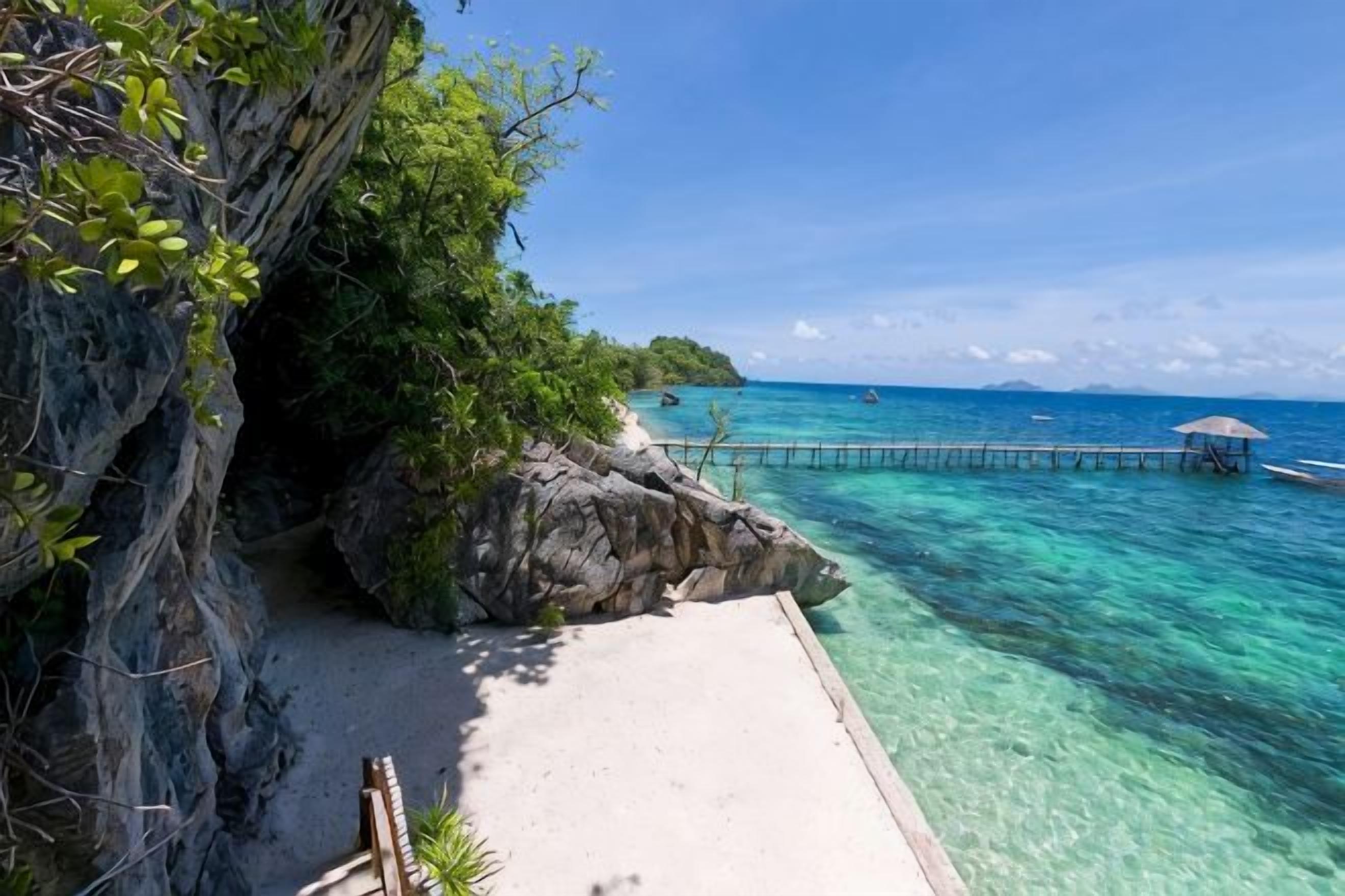 A coastal scene with clear turquoise water, a rocky outcrop, and a pier extending into the sea.
