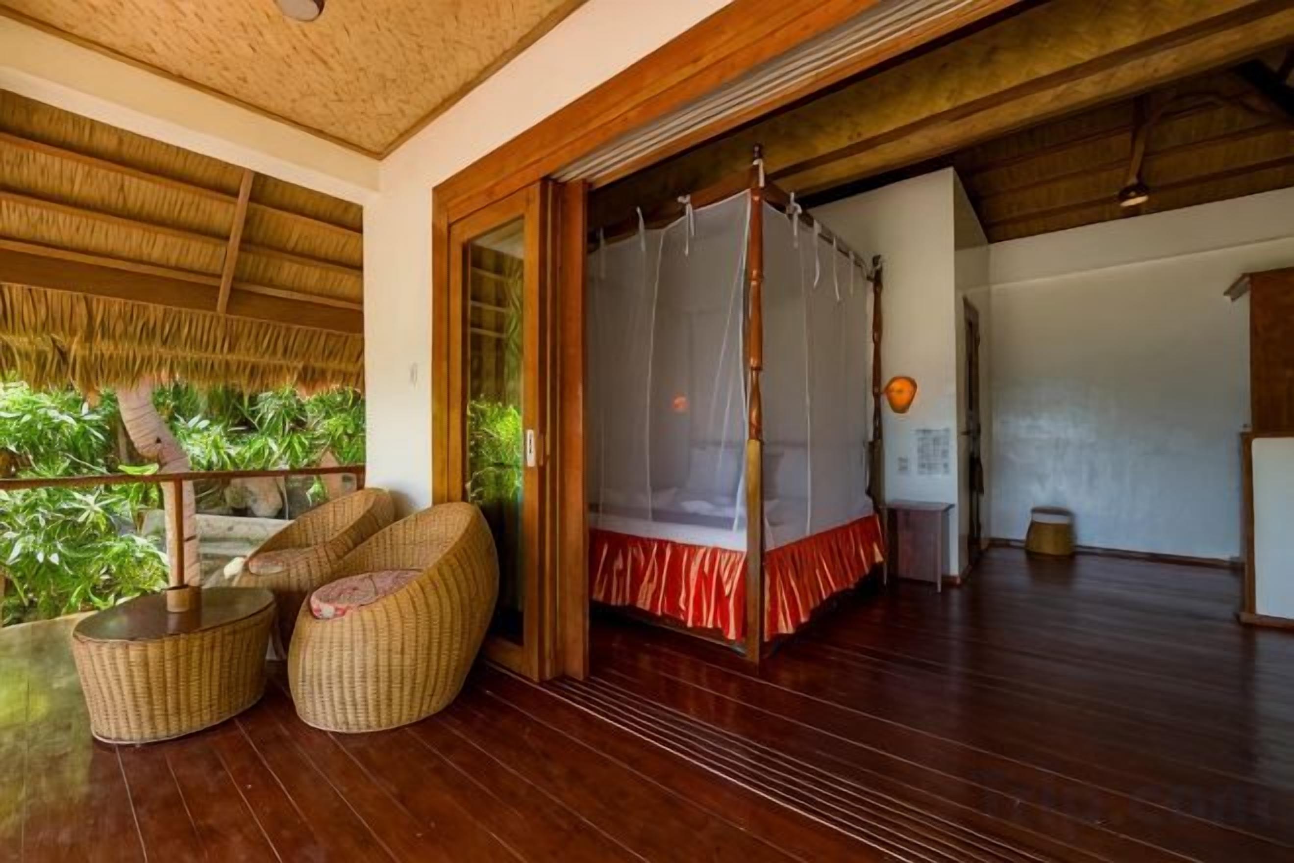 A room with a wooden floor, a bed with a red bedspread, a mosquito net, wicker furniture, and a view of greenery outside.