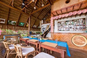 Un restaurante con mesas y sillas de madera, una zona de bar con botellas y un techo de paja.